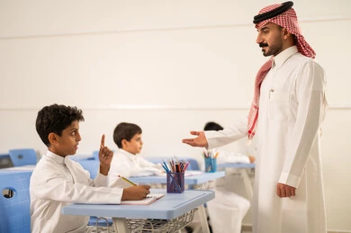 تعابير وايماءات الحماس لمعرفة الجواب الصحيح، طالب عربي خليجي سعودي مبتسم يرتدي الثوب التقليدي يرفع يديه للإجابة، التفاعل والمشاركة داخل الفصل الدراسي، التربية والتعليم بالمناهج المقررة للطلاب في المملكة العربية السعودية