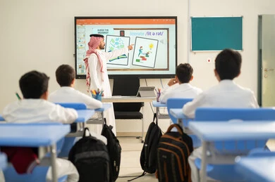 The teaching methods in the schools of the Kingdom have evolved, benefiting from advanced technologies in learning and studying, developed curricula, and e-learning. Two Saudi Gulf Arab students are sitting in their seats, and in front of them is a Saudi teacher using the blackboard to explain.