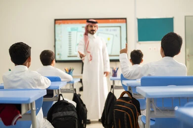 طالب عربي خليجي سعودي مبتسم يرتدي الثوب التقليدي يرفع يديه للإجابة، التفاعل والمشاركة داخل الفصل الدراسي، التربية والتعليم بالمناهج المقررة للطلاب في المملكة العربية السعودية، تعابير وايماءات الحماس لمعرفة الجواب الصحيح