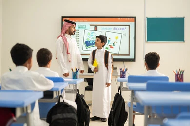 أهداف التربية والتعليم في المملكة العربية السعودية، طلاب عربيين خليجيين سعوديين يجلسون على المقاعد داخل الصف وينتبهون للدرس بايماءات التركيز، تنمية المهارات الإبداعية، مفهوم الاجتهاد والتفوق، مدرس سعودي يقف بجانب طالب ينظر بايماءات السعاده،موسم العودة إلى المدارس