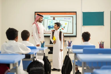 Back to school season, the goals of education in the Kingdom of Saudi Arabia, Arab Gulf Saudi students sitting at their desks in class and paying attention to the lesson with gestures of focus, developing creative skills, the concept of diligence and excellence, a Saudi teacher standing next to a student looking with gestures of happiness.