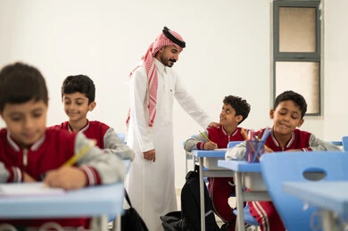 The quality of education in Saudi Arabia, a Gulf Arab teacher wearing traditional attire and a shemagh discussing lessons with his students, a group of Gulf Arab Saudi students in school uniforms completing their assignments, active participation of students with teachers, education and teaching with the prescribed curricula for students, friendly dialogues between the teacher and students.