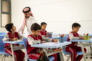 The quality of education in Saudi Arabia, a Gulf Arab teacher wearing traditional attire and a shemagh discussing lessons with his students, a group of Gulf Arab Saudi students in school uniforms completing their assignments, active participation of students with teachers, education and teaching with the prescribed curricula for students, friendly dialogues between the teacher and students.