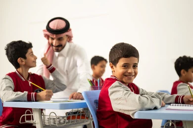 A Gulf Arab Saudi teacher wearing traditional attire and a shemagh is discussing lessons with his students. A group of Arab Gulf Saudi students in school uniforms are doing their homework. There is active participation from the students with the teachers, education and teaching with the prescribed curricula for students, the quality of education in Saudi Arabia, and friendly dialogues between the teacher and the students.