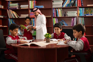 Enjoying reading school books, doing homework, a Gulf Arab Saudi teacher helping his students, friendly dialogues between the teacher and the student, Gulf Arab Saudi students wearing school uniforms sitting in the school library with their teacher, education in the Kingdom of Saudi Arabia.