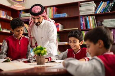 الحوارات الودية بين المعلم والطالب ، طلاب عربيين سعوديين خليجيين يرتدون الزي المدرسي يجلسون في المكتبة المدرسية برفقة معلمهم ، الاستمتاع بقراءة الكتب المدرسية، تأدية الواجبات المدرسية، معلم عربي خليجي سعودي يقوم بمساعده طلابه، التعليم في المملكة العربية السعودية