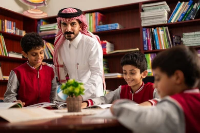 A Gulf Arab Saudi teacher is helping his students, friendly dialogues between the teacher and the student, Arab Saudi Gulf students wearing school uniforms sitting in the school library with their teacher, enjoying reading school books, doing homework, education in the Kingdom of Saudi Arabia.