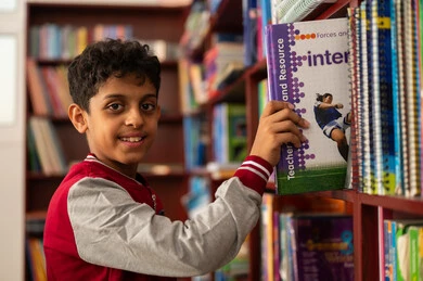 طالب عربي خليجي سعودي يرتدي الزي المدرسي يقف بجوار مكتبة خشبية فى المدرسة ، مفهوم القراءه والثقافه والاستطلاع, مدارس أكاديميه في المملكة العربية السعودية ، بناء مستقبل مشرق للمجتمع، بيئه مدرسيه تعليميه متطوره ، ممارسة هوايه المطالعة، الاستمتاع بقراءة الكتب المدرسية