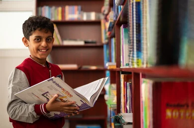 The concept of reading, culture, and exploration, academic schools in the Kingdom of Saudi Arabia, building a bright future for the community, a developed educational school environment, practicing the hobby of reading, an Arab Gulf Saudi student wearing a school uniform standing next to a wooden library in the school, enjoying reading school books.