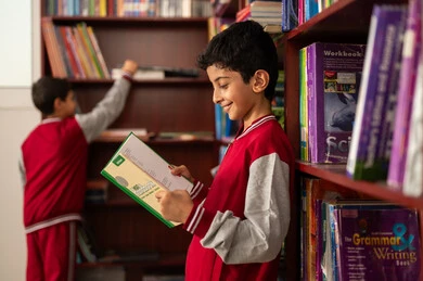 Academic schools in the Kingdom of Saudi Arabia, the concept of reading, culture, and exploration, building a bright future for the community, a developed educational school environment, practicing the hobby of reading, an Arab Gulf Saudi student wearing a school uniform standing next to a wooden library in the school, enjoying reading school books.
