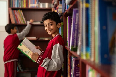 The concept of reading, culture, and exploration, academic schools in the Kingdom of Saudi Arabia, building a bright future for the community, a developed educational school environment, practicing the hobby of reading, an Arab Gulf Saudi student wearing a school uniform standing next to a wooden library in the school, enjoying reading school books.