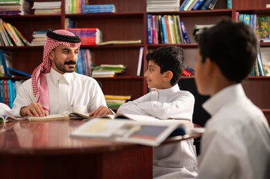 الاستمتاع بقراءة الكتب المدرسية، تأدية الواجبات المدرسية، الحوارات الودية بين المعلم والطالب ، طلاب عربيين سعوديين خليجيين يرتدون الزي التقليدي يجلسون في المكتبة المدرسية برفقة معلمهم ،التعليم في المملكة العربية السعودية