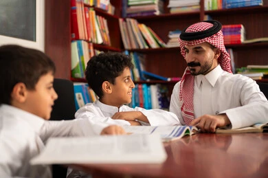 تأدية الواجبات المدرسية، الحوارات الودية بين المعلم والطالب ، طلاب عربيين سعوديين خليجيين يرتدون الزي التقليدي يجلسون في المكتبة المدرسية برفقة معلمهم ، الاستمتاع بقراءة الكتب المدرسية، التعليم في المملكة العربية السعودية