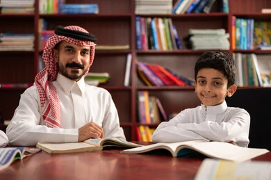 Friendly dialogues between the teacher and the student, a Saudi Gulf Arab student sitting in the school library with their teacher, enjoying reading school books, completing school assignments, education in the Kingdom of Saudi Arabia.
