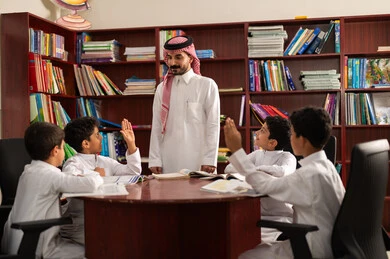 Two Arab Saudi Gulf students are sitting in the school library with their teacher, enjoying reading textbooks, doing homework, and having friendly dialogues between the teacher and the student, education in the Kingdom of Saudi Arabia.