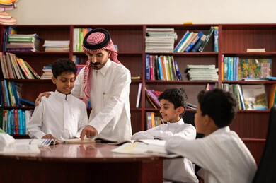 الحوارات الودية بين المعلم والطالب ، طلاب عربيين سعوديين خليجيين يجلسون في المكتبة المدرسية برفقة معلمهم ، الاستمتاع بقراءة الكتب المدرسية، تأدية الواجبات المدرسية، التعليم في المملكة العربية السعودية