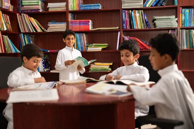 A developed educational school environment, the concept of self-learning, practicing the hobby of reading, a group of Arab Gulf Saudi students wearing traditional attire sitting in the school library, enjoying reading school books, education in the Kingdom of Saudi Arabia.