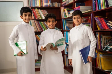 Practicing the hobby of reading, the concept of education and culture, a group of Arab Gulf Saudi students wearing traditional attire standing next to a wooden library in the school library, enjoying reading school books, education in the Kingdom of Saudi Arabia.