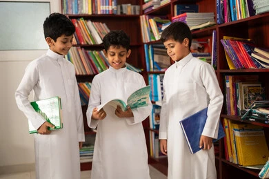 The concept of education and culture, practicing the hobby of reading, a group of Arab Gulf Saudi students wearing traditional attire standing next to a wooden library in the school library, enjoying reading school books, education in the Kingdom of Saudi Arabia.