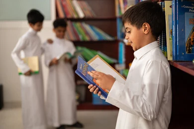 الاستمتاع بقراءة الكتب المدرسية،  ممارسة الهوايات المفضلة ، طالب عربي سعودي يرتدي الزي التقليدي يقف بجوار مكتبة خشبية في المكتبة المدرسية، التعليم في المملكة العربية السعودية