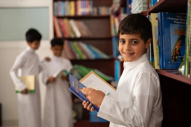 Education in the Kingdom of Saudi Arabia, practicing favorite hobbies, a Saudi Arab student wearing traditional attire standing next to a wooden library in the school library, enjoying reading school books.