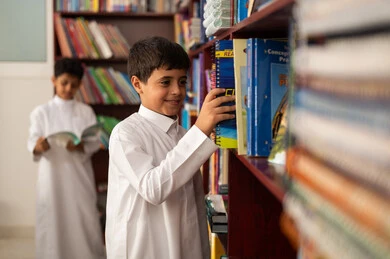 A Saudi Arabian Arab student wearing traditional attire stands next to a wooden bookshelf in the school library, enjoying reading school books, practicing favorite hobbies, education in the Kingdom of Saudi Arabia.