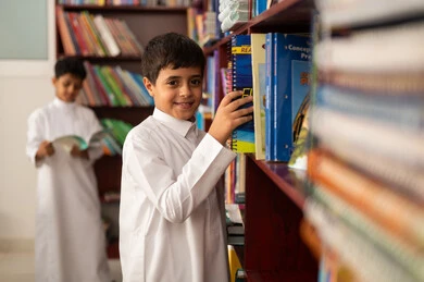 ممارسة الهوايات المفضلة ، طالب عربي سعودي يرتدي الزي التقليدي يقف بجوار مكتبة خشبية في المكتبة المدرسية، الاستمتاع بقراءة الكتب المدرسية، التعليم في المملكة العربية السعودية