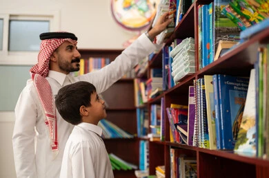 طالب عربي سعودي يرتدي الزي التقليدي يقف بجوار مكتبة خشبية في المكتبة المدرسية برفقة معلمه ، الحوارات الودية بين المعلم والطالب، الاستمتاع بقراءة الكتب المدرسية، ممارسة الهوايات المفضلة ، التعليم في المملكة العربية السعودية