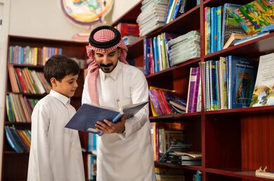 ممارسة هواية المطالعة ، الحوارات الودية بين المعلم والطالب، الاستمتاع بقراءة الكتب المدرسية، طالب عربي سعودي يرتدي الزي التقليدي يقف بجوار مكتبة خشبية في المكتبة المدرسية برفقة معلمه ، التعليم في المملكة العربية السعودية