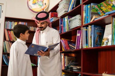 الاستمتاع بقراءة الكتب المدرسية، الحوارات الودية بين المعلم والطالب، ممارسة الهوايات المفضلة ، طالب عربي سعودي يرتدي الزي التقليدي يقف بجوار مكتبة خشبية في المكتبة المدرسية برفقة معلمه ، التعليم في المملكة العربية السعودية