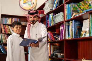 الحوارات الودية بين المعلم والطالب، الاستمتاع بقراءة الكتب المدرسية، ممارسة الهوايات المفضلة ، طالب عربي سعودي يرتدي الزي التقليدي يقف بجوار مكتبة خشبية في المكتبة المدرسية برفقة معلمه ، التعليم في المملكة العربية السعودية