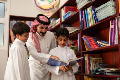 مفهوم الود والالفه ،مساعدة المعلم لطلابه، الاستمتاع بقراءة الكتب المدرسية، ممارسة الهوايات المفضلة ، مجموعة من طلاب عربيين خليجيين سعوديين يرتدون الزي التقليدي يقفون بجوار مكتبة خشبية في المكتبة المدرسية برفقة معلمه ، التعليم في المملكة العربية السعودية