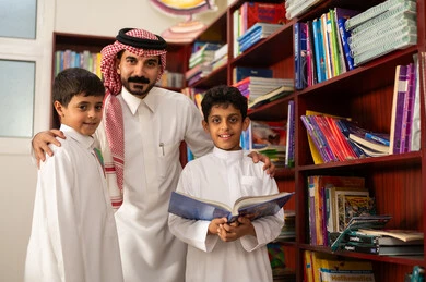التعليم في المملكة العربية السعودية، مساعدة المعلم لطلابه، مفهوم الود والالفه ،الاستمتاع بقراءة الكتب المدرسية، ممارسة الهوايات المفضلة ، مجموعة من طلاب عربيين خليجيين سعوديين يرتدون الزي التقليدي يقفون بجوار مكتبة خشبية في المكتبة المدرسية برفقة معلمه