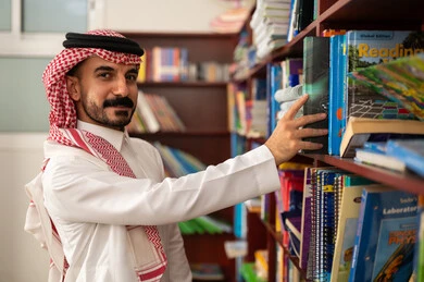 A Saudi Arabic teacher is taking a book from a wooden shelf in the school library, practicing the hobby of reading, education in the Kingdom of Saudi Arabia, enjoying reading school books.
