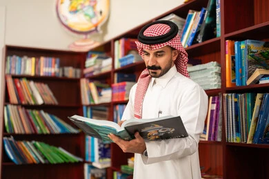 Enjoying reading school books, education in the Kingdom of Saudi Arabia, practicing favorite hobbies, a Saudi Arab teacher standing next to a wooden library in the school library.