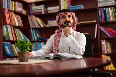 تعابير التفكير والتأمل، مفهوم المطالعة والثقافة ، التعليم في المملكة العربية السعودية، ممارسة الهوايات المفضلة ، معلم عربي سعودي يرتدي الزى التقليدي والشماغ يجلس في المكتبة المدرسية، الاستمتاع بقراءة الكتب المفيدة