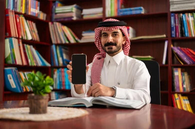 عرض شاشة سوداء فارغة، الدردشة عبر الانترنت ، اجراء مكالمه هاتفية  ، التعليم في المملكة العربية السعودية، ممارسة الهوايات المفضلة ، معلم عربي سعودي يرتدي الزى التقليدي والشماغ يجلس في المكتبة المدرسية يمسك بيده الجوال ، الاستمتاع بقراءة الكتب المفيدة