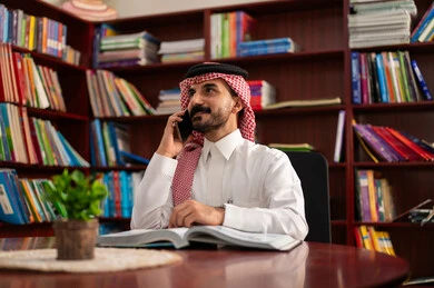 اجراء مكالمه هاتفية  ، التعليم في المملكة العربية السعودية، استخدام التقنيات الحديثة، الدردشة عبر الانترنت ،ممارسة الهوايات المفضلة ، معلم عربي سعودي يرتدي الزى التقليدي والشماغ يجلس في المكتبة المدرسية يمسك بيده الجوال ، الاستمتاع بقراءة الكتب المفيدة