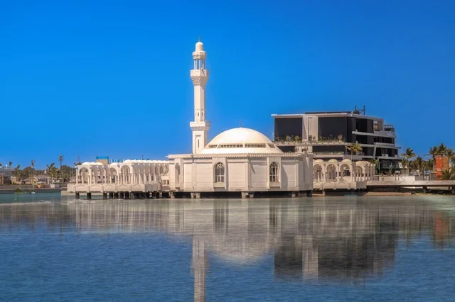 Mosques, hotels, and parks on the waterfront, a charming daytime scene of Jeddah's beach and its distinctive landmarks, the meeting of the clear blue waters of the Red Sea with the vast horizon, in a tranquil atmosphere that reflects the beauty of the city of Jeddah and the splendor of its coastal nature.