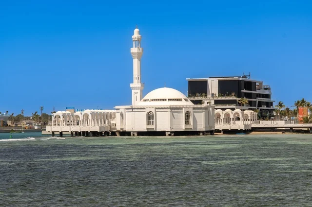 التقاء مياه البحر الأحمر الزرقاء الصافية مع الأفق الواسع، مشهد نهاري ساحر لشاطئ جدة ومعالمها المميزة ،  مساجد وفنادق ومنتزهات على الواجهة البحرية، أجواء هادئة تعكس جمال مدينة جدة وروعة طبيعتها الساحلية.