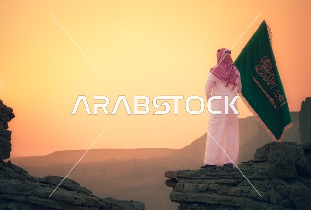 رفع علم المملكة العربية السعودية وسط الصحراء، راية التوحيد وشعار الوطن، الاحتفال بالمناسبات والأعياد الوطنية، يوم العلم 11 مارس، اليوم الوطني السعودي 23 سبتمبر، صورة من الخلف لرجل عربي خليجي سعودي يرتدي الشماغ والثوب التقليدي يعتز بتراب الوطن