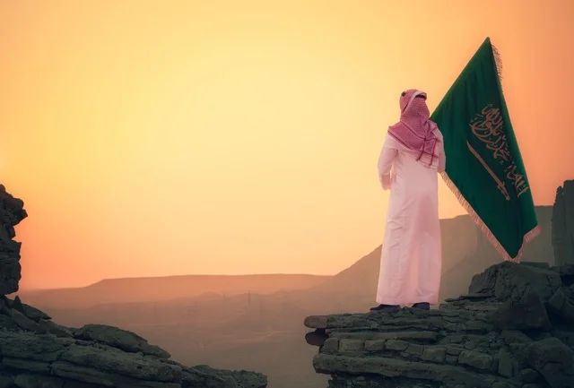 The flag of the Kingdom of Saudi Arabia is raised in the middle of the desert, the banner of monotheism and the emblem of the homeland, celebrating national occasions and holidays, Flag Day on March 11, Saudi National Day on September 23, a picture from behind of an Arab Gulf Saudi man wearing the ghutrah and traditional thobe, proud of the soil of the homeland.
