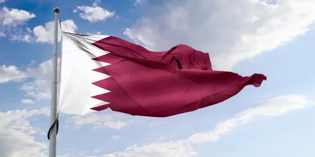 The flag of the State of Qatar flutters in the blue sky, celebrating Qatar National Day, the Islamic Arts Museum in the Qatari capital Doha, famous tourist attractions during the day, traditional mud buildings.