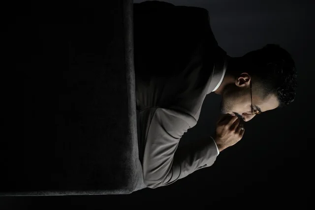 رجل عربي أنيق يرتدي بدلة رسمية رمادية ونظارات طبية، يجلس بتأمل ووقار في إضاءة احترافية سوداء، مشهد يعكس الثقة، الفخامة، والحياة المهنية المرموقة