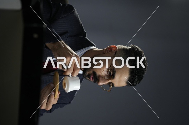 رجل عربي أنيق يرتدي بدلة رسمية ونظارات طبية يحتسي القهوة العربية في أجواء هادئة، تعبير عن الفخامة واللحظة الخاصة بالاسترخاء أثناء العمل 