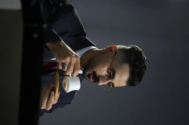 رجل عربي أنيق يرتدي بدلة رسمية ونظارات طبية يحتسي القهوة العربية في أجواء هادئة، تعبير عن الفخامة واللحظة الخاصة بالاسترخاء أثناء العمل 