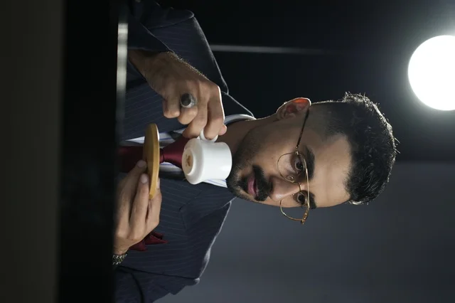 صورة تعبر عن الفخامة والنجاح ورجال الأعمال ، رجل أنيق يرتدي بدلة رسمية وربطة عنق حمراء يشرب القهوة في فنجان صغير، يبدو عليه التركيز والهدوء أثناء الاستمتاع بالمشروب الساخن