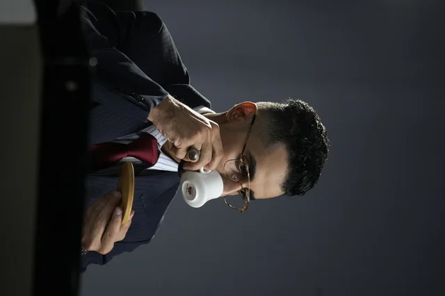 An elegant Arab Gulf man wearing a classic formal suit, holding a cup of coffee with a small wooden saucer, looking with focus and harmony. The image reflects refined taste, self-confidence, and a business atmosphere