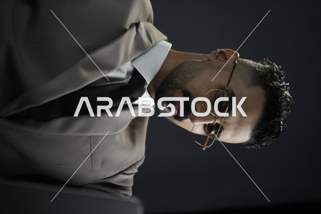 رجل عربي خليجي يرتدي بدلة رسمية داكنة اللون مع ربطة عنق ونظارات، يجلس على مكتب حديث في بيئة عمل مكتبية راقية