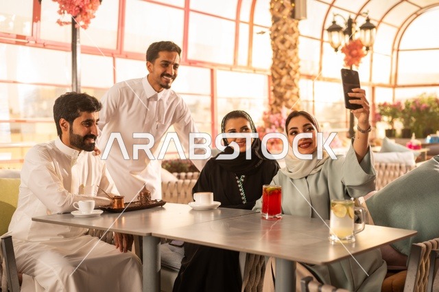 استخدام اتكنولوجيا والتقنيات الحديثة، توثيق اللحظات السعيدة من خلال التقاط الصور التذكارية، إمرأة عربية خليجية سعودية ترتدي العباءة والحجاب تجلس على الأريكة مع أصدقائها بإحدى المقاهي وتستخدم الهاتف، مشاركة المشروبات الباردة والحلويات